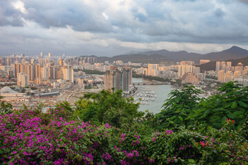 Panoramic view of Sanya, Hainan Island, China. Lush tropical foliage and purple bougainvillea frame on foreground, marina with moored boats, high-rise buildings and distant mountains on background.