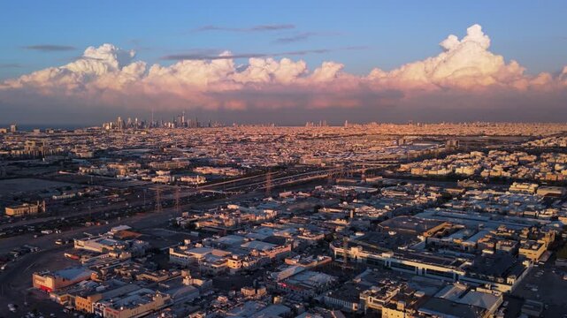 Wide drone view of Kuwait City at golden hour: distant skyline on the Gulf coast, towering cumulonimbus, vast residential grid, highways and power pylons; warm sunset glow and clear visibility.