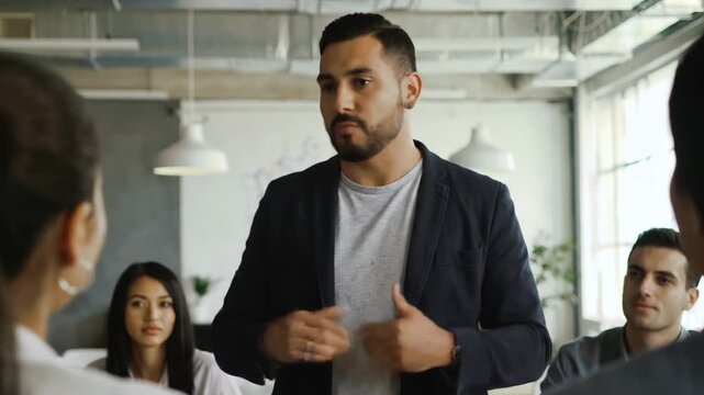Confident Hispanic man leading a business meeting, talking to a diverse group of people in modern office. Teamwork and communication.