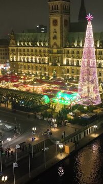 Aerial footage of Hamburg Christmas Market in front of the historic City Hall, illuminated by festive lights. Aerial view of towering Christmas tree near Hamburg Christmas Market.