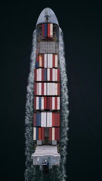 Top-down aerial view of container ship cutting through dark water, leaving white wake. Cargo vessel in motion, symbol of global shipping, logistics, trade and maritime transportation