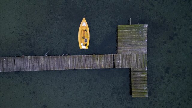Top-down aerial view of small yellow boat moored at wooden pier over clear water. Minimal coastal scene with calm atmosphere, ideal for travel, nature, leisure and nautical themes