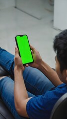 Young man holding green screen phone while sitting on couch