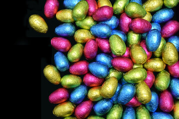 Pile or group of multi colored and different sizes of colourful foil wrapped chocolate easter eggs in pink, blue, yellow and lime green against a black background with copy space.