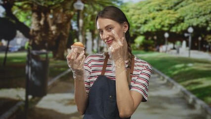 Woman holding muffin with gloved hand and pointing finger to cheek while wearing apron and striped shirt on street path by trees and lamp posts; friendly service.