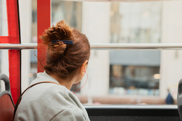 A woman with her brown hair tied in a bun looks out the window of a red bus while wearing red wired earphones © Contentino