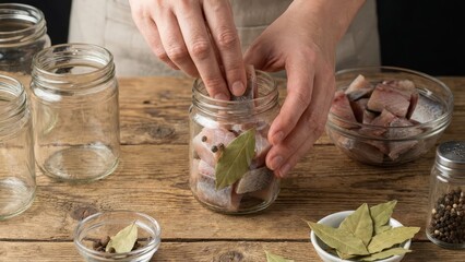 Hands arranging fresh fish pieces with bay leaves and peppercorns into a glass mason jar on a rustic wooden table, embodying homemade food preservation and culinary tradition
