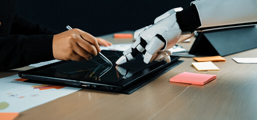 A robot arm interacts with a human hand on a tablet, showcasing a collaborative effort in a modern workspace. This image highlights innovation, technology, and teamwork.Noogenesis
