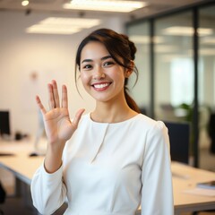 A cheerful young Asian businesswoman smiles confidently while holding up her hand in a greeting or stopping gesture inside a modern, bright, blurred office environment.