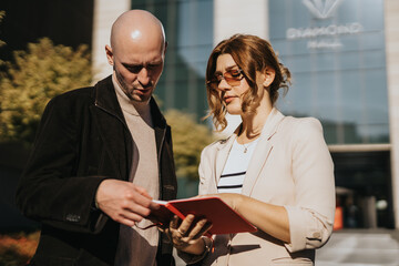Two business colleagues review a red notebook outside a contemporary office building. They discuss files and plans in a casual outdoor setting.