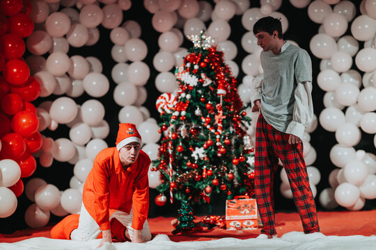 Two men in costumes stand and kneel beside a decorated Christmas tree. A festive scene with balloons and gifts.