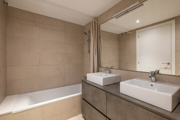 Modern bathroom with a bathtub, two sinks, and a large mirror. Neutral color scheme with tiled walls and a sleek design.