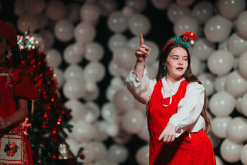 A woman in a red dress and white blouse performs on a stage decorated with white balloons. The festive scene captures a Christmas theater show with dancers and holiday cheer.