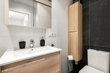 Modern bathroom with a sleek design. Features a white sink, wooden cabinetry, a mirror, and a toilet. The walls are tiled in black and white.