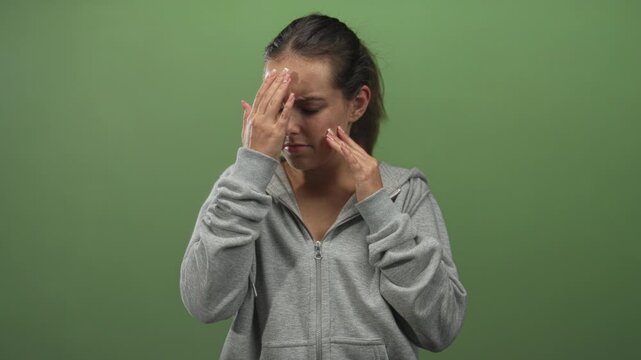 Young woman touching her temples with both hands, face strained in studio with green backdrop; headache pain.