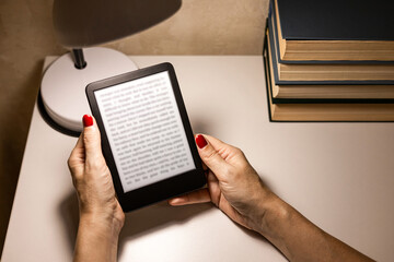  A woman reading an e-book at home. reading an e-book by lamplight. reading a book at the table. woman holding e-book. hand holding ereader. e-ink book.