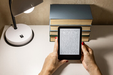  A woman reading an e-book at home. reading an e-book by lamplight. reading a book at the table. woman holding e-book. hand holding ereader. e-ink book. an alternative to paper books