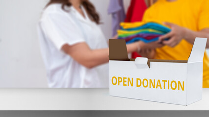 White box labeled "OPEN DONATION" sitting on a white table with people in the background donating colorful clothes. Charitable activities, volunteering, and donation drives