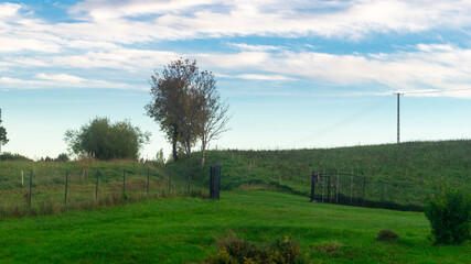 A yard with green trimmed grass and blue sky. A road with green grass leading to the yard against the backdrop of a hill