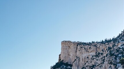 Majestic rock cliffs rise against a clear blue sky, dotted with evergreen trees.