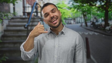Man wearing striped shirt showing call gesture with hand on street scene; friendly invitation social connection.