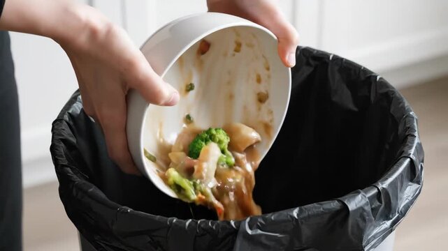Tossing a bowl of cooked vegetables into a trash can, highlighting food waste, Discarding leftover cooked vegetables into a black garbage bag
