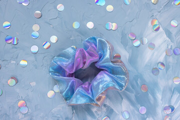 Iridescent satin hair scrunchie on soft blue background with holographic confetti, modern fashion accessory flat lay with playful aesthetic mood