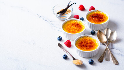 Creamy cr&egrave;me br&ucirc;l&eacute;e dessert in ramekins with caramelized sugar topping, surrounded by fresh berries, vanilla pods, and spoons on a white marble surface, culinary master class, food blog, copy space
