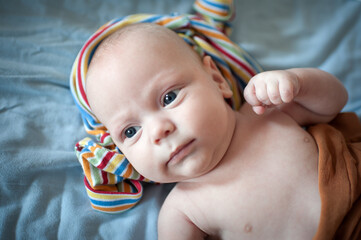 A baby is lying on a soft blanket. The baby has a cloth with bright stripes draped on the head. The baby looks towards the camera with wide eyes.