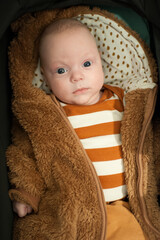A baby is inside a stroller wearing a warm brown coat and a striped shirt. The baby looks calm and curious while resting outdoors