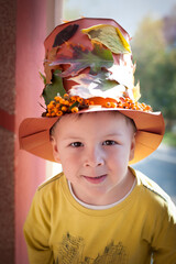 A child stands outside a house wearing a colorful hat made of leaves and flowers. The sun is shining, and the leaves show bright autumn colors.