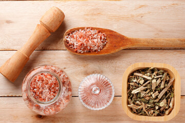 Himalayan salt in a wooden spoon, accompanied by a grinder, glass jar, and container with dried herbs on a light wood surface. Top view.