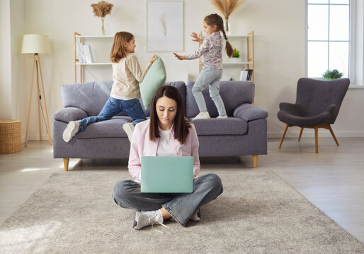 Portrait of young mother sitting on floor in living room at home and working on laptop computer while her noisy little kids playing pillow fight on sofa in background. Family and motherhood concept.