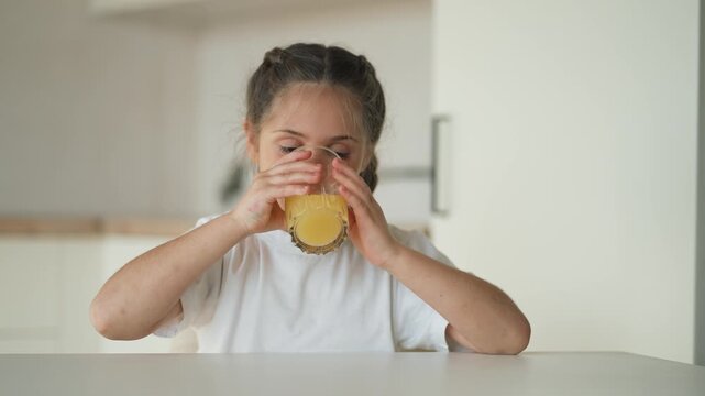 Girl sits at kitchen table smiling while holding glass of orange juice for breakfast in modern kitchen setting child enjoys healthy drink and smiles toward glass with content expression and joy