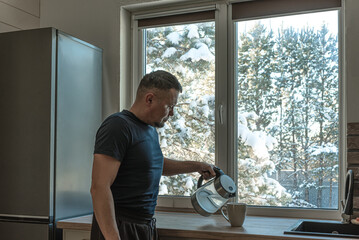  Senior man pours water from kettle to mug near winter window