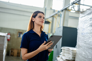 Female factory worker using digital tablet in industrial warehouse, monitoring production and inventory management. Concept of manufacturing process, smart factory, technology, efficiency