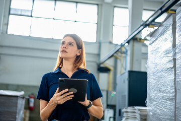Female factory worker using digital tablet in industrial warehouse, monitoring production and inventory management. Concept of manufacturing process, smart factory, technology, efficiency