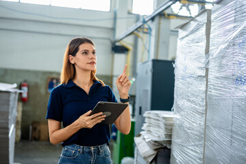 Female factory worker using digital tablet in industrial warehouse, monitoring production and inventory management. Concept of manufacturing process, smart factory, technology, efficiency