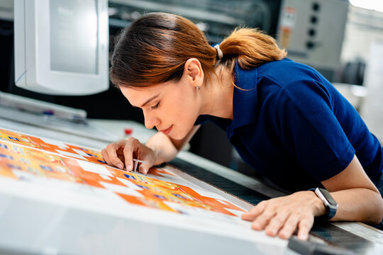 Female industrial worker inspecting printed materials on production table, checking color accuracy and print quality in modern factory. Concept of quality control, printing process, precision work.