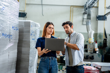 Two professional factory workers use a laptop to manage inventory and production workflow in a printing plant, ensuring high-tech manufacturing quality control and industrial warehouse efficiency