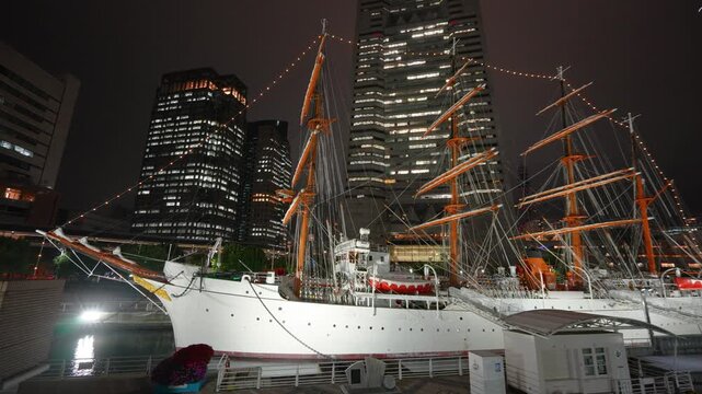 Historic Nippon Maru sailing ship illuminated at night in Yokohama. The scene shows modern city buildings in Minato Mirai during the autumn season.