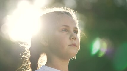 Young girl holds thoughtful gaze toward bright sunlight with backlight halo around braid and face in outdoor nature portrait of child in green environment showing calm expression and gentle light