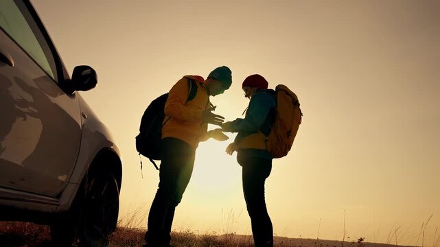Hikers check map at sunset while backpacker and partner adjust pack beside parked car creating warm silhouette against nature field travel roadtrip with backpack and map visible under golden sky