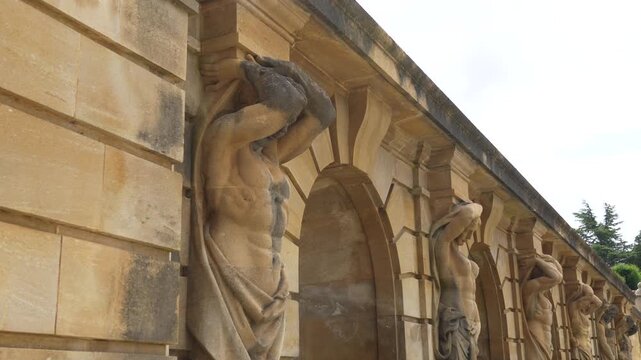 Series of sandstone sculptures supporting arched wall with repeating figures beside stone stairs and green trees.