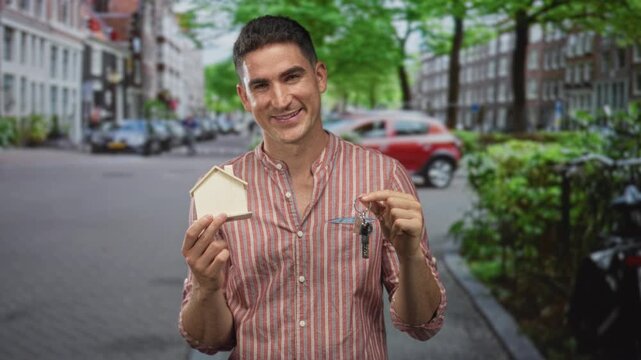 Man holding small wooden house model and keys with both hands on a leafy street in city; homeownership pride.