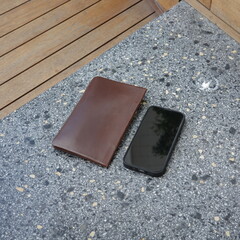 A brown leather wallet and a black smartphone resting on a stone tabletop outdoors.