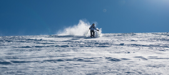 Naklejka premium Skier skiing off piste through fresh powder on a sunny mountain slope, kicking up snow as she descends, creating a dynamic and energetic winter sports action scene under a clear blue sky