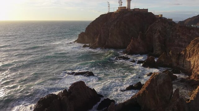 Scenic aerial view of waves crashing against the rocky coast by the Cabo de Gata lighthouse at sunset