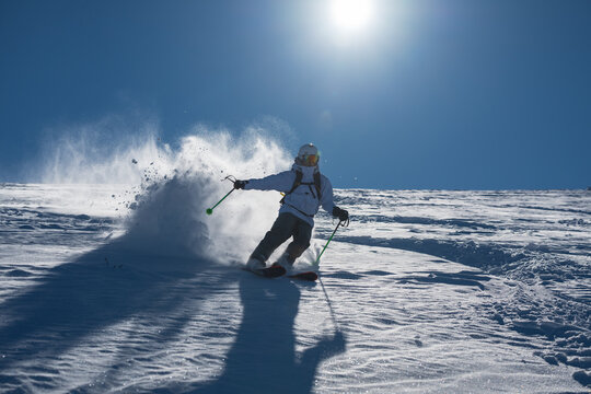Skier skiing off piste through fresh powder on a sunny mountain slope, kicking up snow as she descends, creating a dynamic and energetic winter sports action scene under a clear blue sky