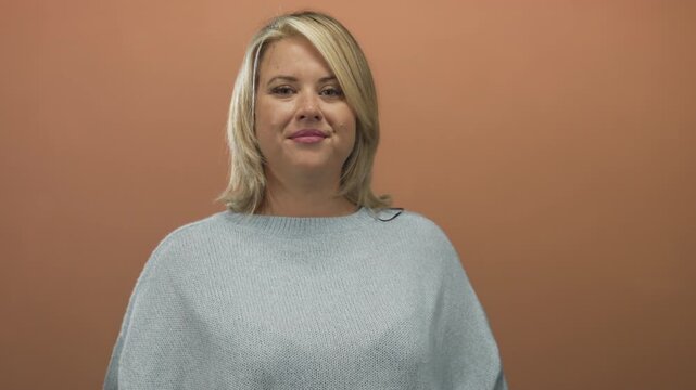 Woman looking at camera while standing in studio wearing a soft loose light blue sweater; confidence.